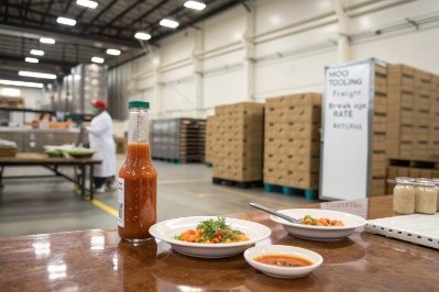 Glass hot sauce bottle on factory table with plated food samples in warehouse setting