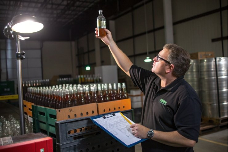 Quality inspector checks amber bottle batch with clipboard under warehouse lighting