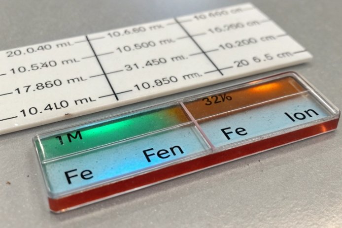Close-up of a colorimetric/indicator slide labeled for iron (Fe), showing a green-to-amber gradient and measurement marks behind it.