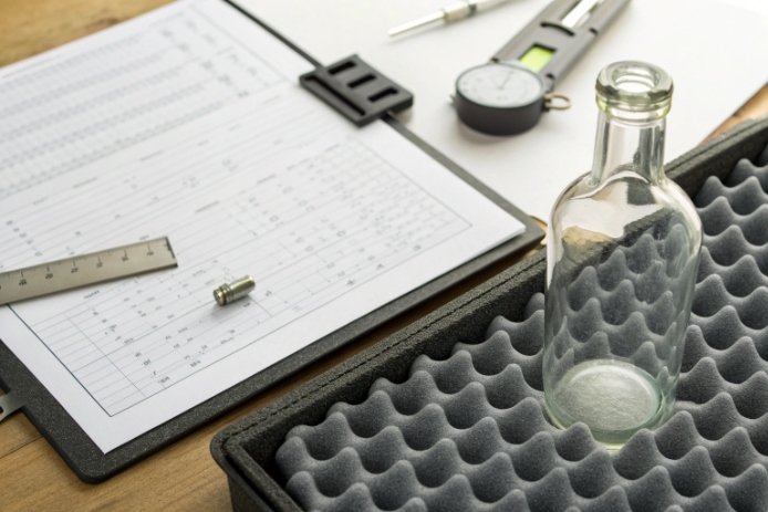 Empty glass bottle in foam tray beside calipers and inspection paperwork for quality control
