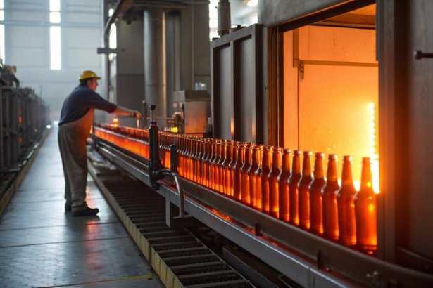 Glowing glass bottles on hot annealing conveyor beside furnace with worker monitoring line