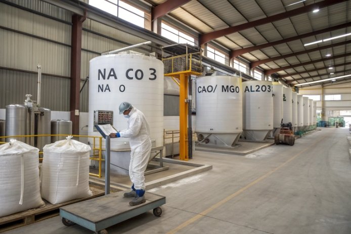 Worker inspecting raw material silos labeled NaCO3, CaO/MgO, Al2O3