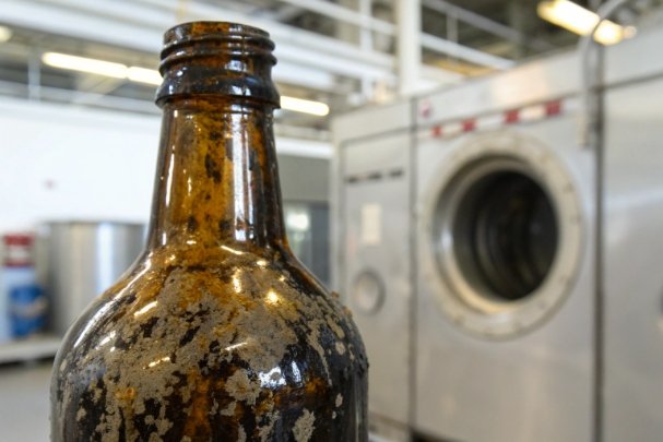Close-up amber glass bottle with heavy residue buildup near industrial washer equipment