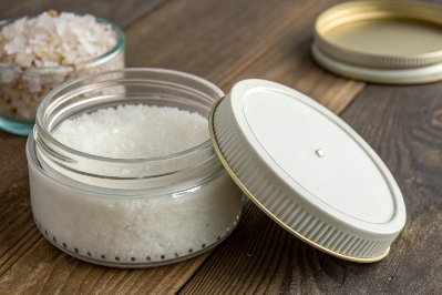 Open glass cosmetic jar filled with bath salts beside metal lid on wooden table