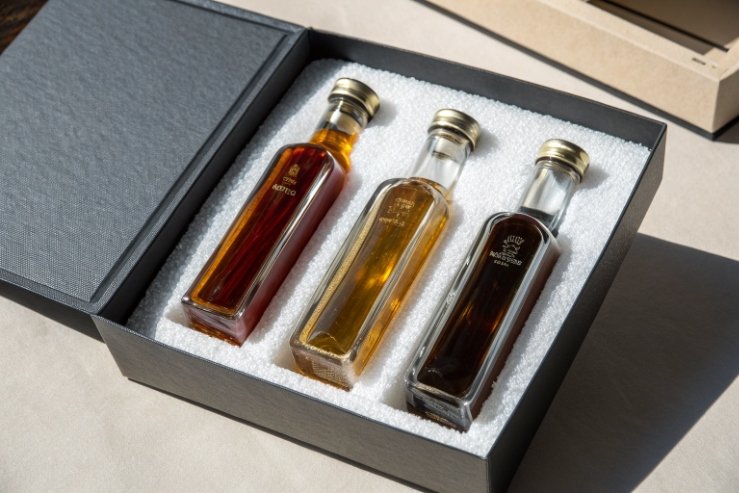 Premium gift set: three small rectangular glass bottles of amber and dark liquids nestled in a foam insert inside a black presentation box