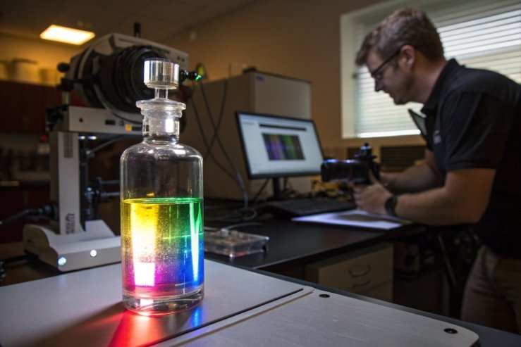 Quality lab test bottle with rainbow liquid, technician recording results in background.
