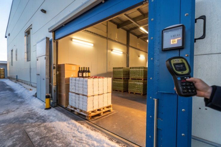 Cold storage loading dock with palletized bottles and handheld temperature scanner check.