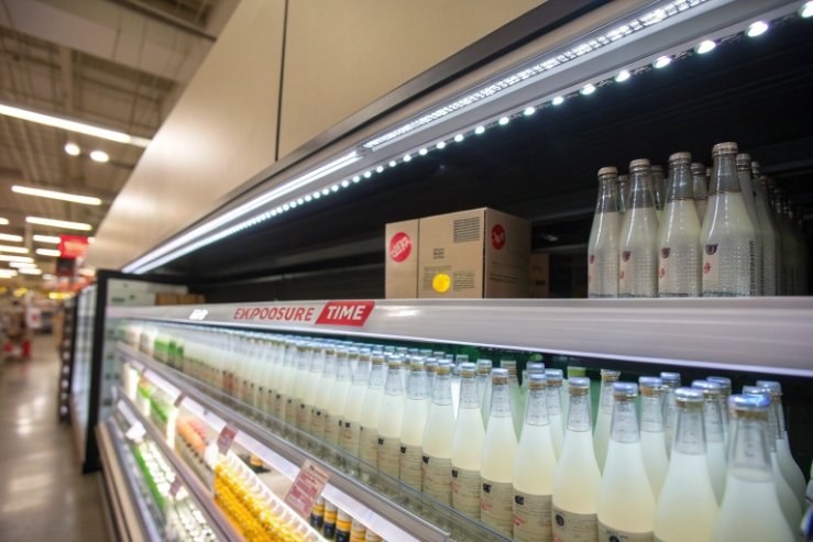 Refrigerated retail shelf with glass beverage bottles under LED lighting highlighting exposure time risk