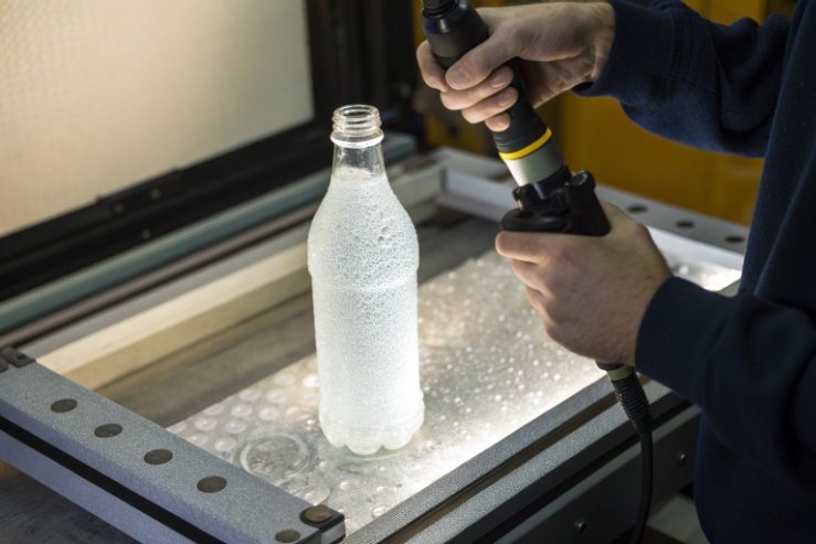 Technician using handheld tool to inspect frosted bottle surface in QC workstation