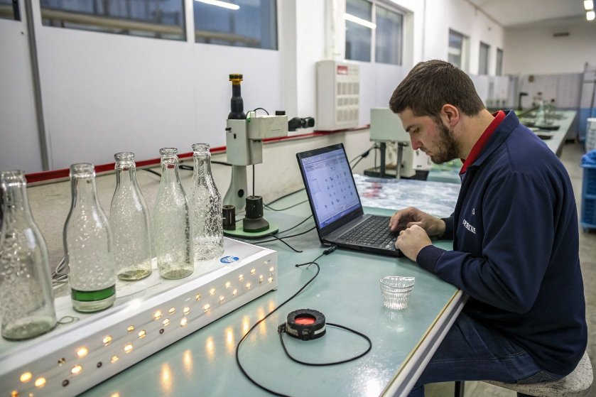 Technician recording glass bottle inspection data on laptop beside illuminated sample bottles