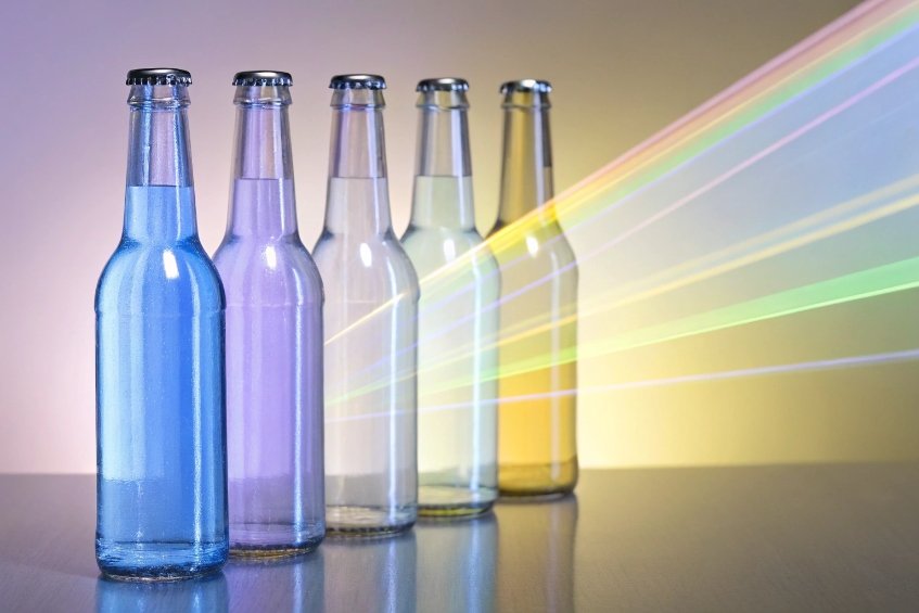 Row of five capped glass bottles filled with faintly tinted liquids, shifting from blue to purple to clear to pale yellow, as diagonal bands of rainbow-colored light shine through them against a soft gradient background