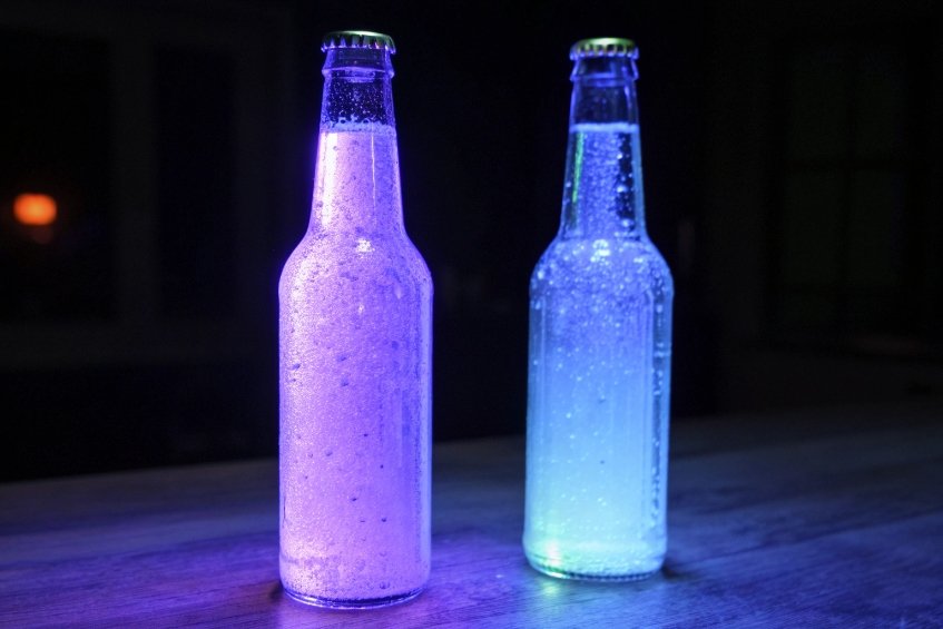 Two long-neck glass bottles on a wooden surface in a dark room, their contents glowing brightly in neon purple and blue with condensation on the glass, creating a dramatic illuminated effect