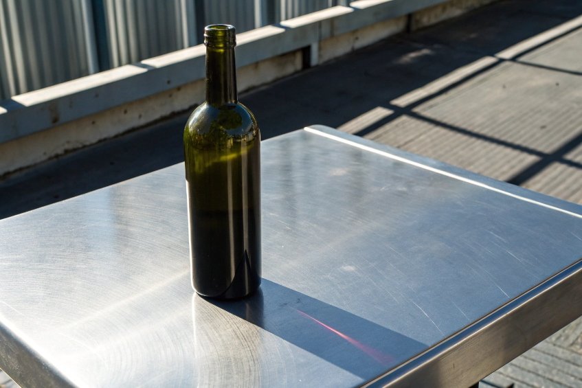 Single green glass wine bottle standing on outdoor stainless steel table in sunlight